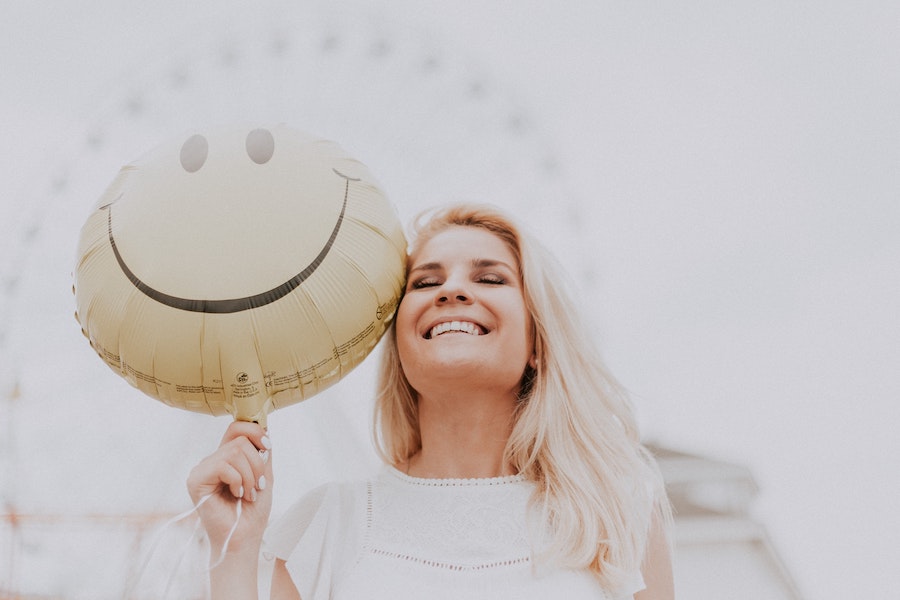 Blonde woman smiles as she holds a smiley balloon by a Ferris wheel