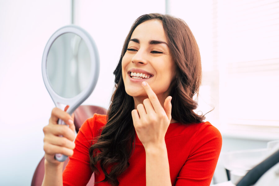 Brunette woman in a red blouse smiles at her teeth while looking at a handheld mirror