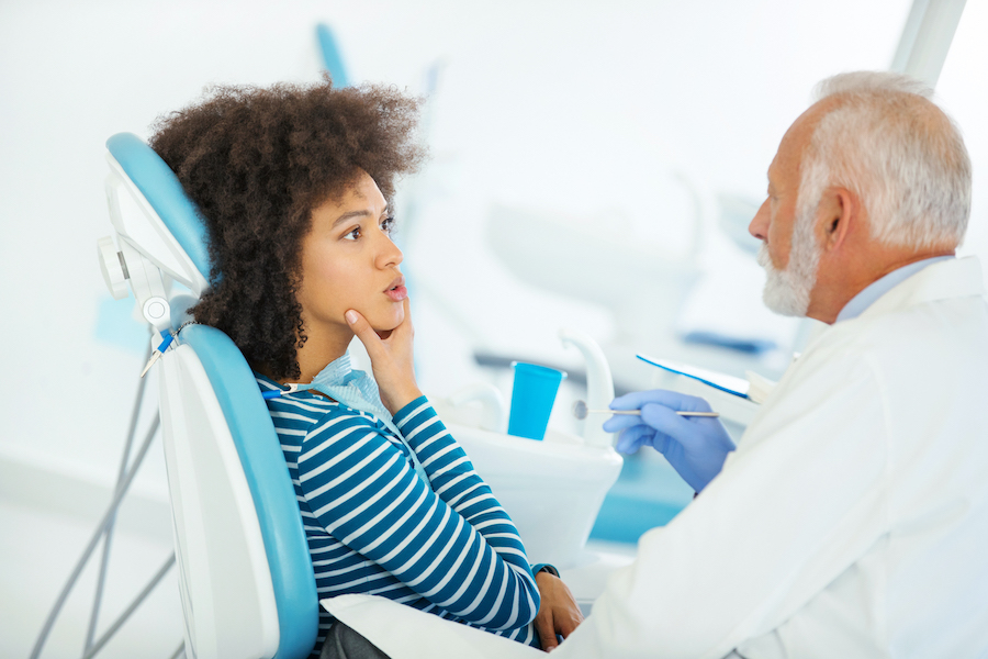 Black woman holds her chin and sits in a dental chair as she talks to her dentist about her dental concern