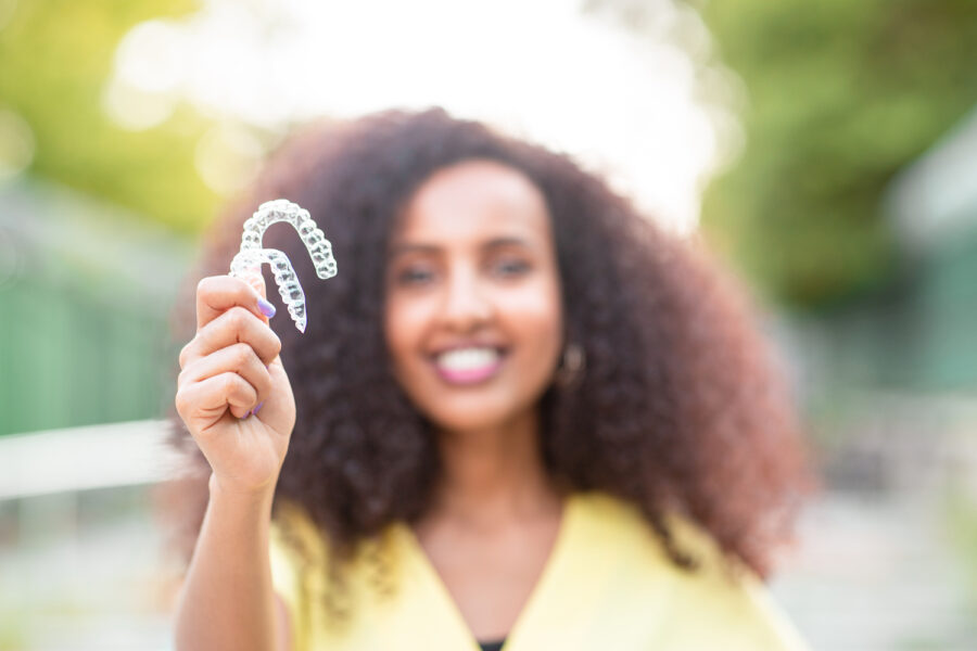 Blurred Black woman smiling in a yellow blouse while holding up her Invisalign clear aligners outside
